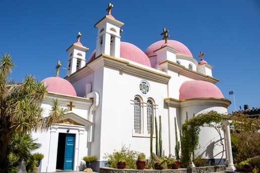 A church with pink domes and white walls is set against a clear blue sky. The structure features several crosses atop its domes and multiple arched windows with decorative designs. In the foreground, there are several tall cacti and other desert plants.