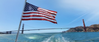 An American flag is waving prominently in the foreground against a backdrop of the Golden Gate Bridge spanning over the blue waters. The sky is clear with a bright blue hue, and the scene is sunlit indicating a clear day.