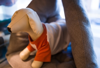 A plush dog mannequin dressed in a small orange and white shirt sits on a soft gray cushion in a cozy setting. The background is softly blurred, suggesting an indoor environment.