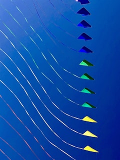 A series of colorful kites are lined in a diagonal pattern against a solid blue sky. The kites vary in shades of blue, green, and yellow, and are attached to thin strings that create wavy lines, adding a sense of movement to the image.