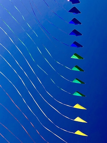 A series of colorful kites are lined in a diagonal pattern against a solid blue sky. The kites vary in shades of blue, green, and yellow, and are attached to thin strings that create wavy lines, adding a sense of movement to the image.