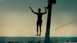 A silhouetted person is balancing on a slackline stretched between two points near a beach or waterfront. The background consists of the ocean, a clear sky, and some distant figures. The scene appears to be taken during a sunset or sunrise, casting a serene ambiance.