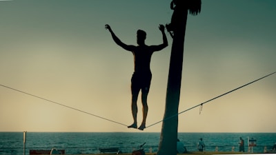 A silhouetted person is balancing on a slackline stretched between two points near a beach or waterfront. The background consists of the ocean, a clear sky, and some distant figures. The scene appears to be taken during a sunset or sunrise, casting a serene ambiance.