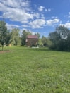 A small wooden cabin with a red roof is surrounded by lush greenery, under a sky filled with scattered white clouds. A group of white ducks is gathered in the middle of the grassy area, which stretches across most of the foreground. Trees frame the background, adding to the serene and pastoral setting.