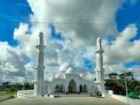 A large white mosque with two tall minarets is set against a vibrant blue sky with scattered white clouds. The mosque features intricate architectural details, including domes and arches, surrounded by lush greenery.