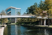 A modern office building with reflective glass windows is situated next to a calm waterway. A pedestrian bridge crosses the water, connecting two sides lined with lush green trees. The sky is clear and blue, adding to the serene atmosphere.