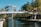 A modern office building with reflective glass windows is situated next to a calm waterway. A pedestrian bridge crosses the water, connecting two sides lined with lush green trees. The sky is clear and blue, adding to the serene atmosphere.