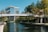 A modern office building with reflective glass windows is situated next to a calm waterway. A pedestrian bridge crosses the water, connecting two sides lined with lush green trees. The sky is clear and blue, adding to the serene atmosphere.
