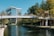 A modern office building with reflective glass windows is situated next to a calm waterway. A pedestrian bridge crosses the water, connecting two sides lined with lush green trees. The sky is clear and blue, adding to the serene atmosphere.