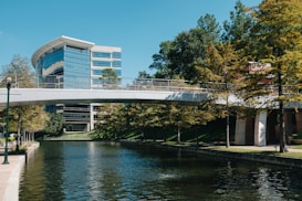 A modern office building with reflective glass windows is situated next to a calm waterway. A pedestrian bridge crosses the water, connecting two sides lined with lush green trees. The sky is clear and blue, adding to the serene atmosphere.