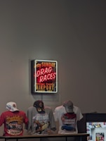 A neon sign advertising drag races at the Orange County Airport in Santa Ana is mounted on the wall. Below the sign, three t-shirts featuring vibrant designs related to cars and motorsport are displayed. Each t-shirt is accessorized with a cap, and there is also a small display case to the side.