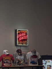 A neon sign advertising drag races at the Orange County Airport in Santa Ana is mounted on the wall. Below the sign, three t-shirts featuring vibrant designs related to cars and motorsport are displayed. Each t-shirt is accessorized with a cap, and there is also a small display case to the side.
