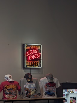 A neon sign advertising drag races at the Orange County Airport in Santa Ana is mounted on the wall. Below the sign, three t-shirts featuring vibrant designs related to cars and motorsport are displayed. Each t-shirt is accessorized with a cap, and there is also a small display case to the side.