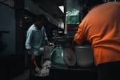Two people are working in a dimly lit room, with one man stirring a pot and the other facing away while operating a machine. A large scale is visible alongside various bags and containers. The setting appears to be industrial or kitchen-like, with metal surfaces and tools around.