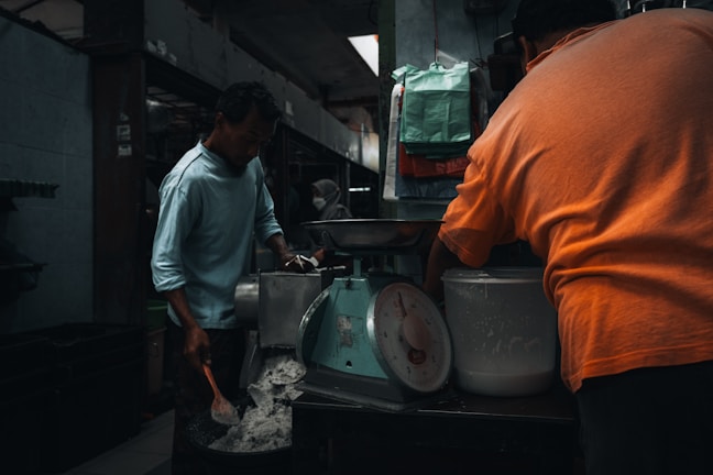 Two people are working in a dimly lit room, with one man stirring a pot and the other facing away while operating a machine. A large scale is visible alongside various bags and containers. The setting appears to be industrial or kitchen-like, with metal surfaces and tools around.