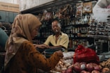 A market scene with a vendor standing behind a counter filled with garlic, onions, and red mesh bags. The vendor, who has a cigarette in his mouth, is engaging with a customer who is wearing a headscarf. Shelves behind the vendor are stocked with various bottles and packets, suggesting a small grocery shop.