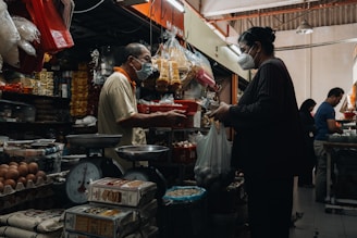 A bustling indoor market scene with a vendor and customer engaged in a transaction. Both individuals are wearing masks. The stall is filled with various goods including eggs, packaged foods, and other groceries. There are old-fashioned weighing scales on the counter and the space is crowded with products.