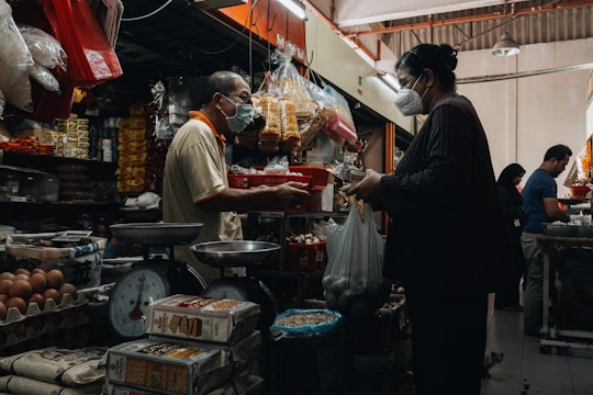 A bustling indoor market scene with a vendor and customer engaged in a transaction. Both individuals are wearing masks. The stall is filled with various goods including eggs, packaged foods, and other groceries. There are old-fashioned weighing scales on the counter and the space is crowded with products.