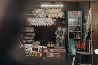 A small kiosk features a variety of packaged snacks and beverages. Hanging displays exhibit several magazines and comic books. A man stands inside on the left, while a child in a blue shirt and striped pants faces the refrigerator filled with drinks on the right.