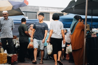A bustling market scene with several people walking among stalls. One person is wearing a blue t-shirt that reads 'Tropical Life' and carrying a plastic bag. Another is wearing a white t-shirt and holding shopping bags. There are tents set up, providing shade in the background, and various goods are on display.