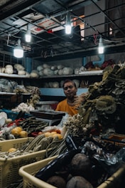 A market scene featuring a vendor surrounded by a variety of vegetables and produce. The setting appears to be a small shop with dim lighting provided by overhead bulbs. Various vegetables such as cucumbers, eggplants, and leafy greens are displayed in baskets and on shelves.