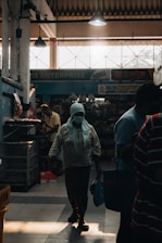 A dimly lit indoor market with a person wearing a headscarf and mask walking through the aisle. The light from outside creates dramatic shadows and silhouettes. Various goods are visible in the background under artificial lighting, and parts of the store signage can be seen.