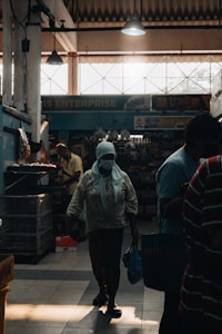 A dimly lit indoor market with a person wearing a headscarf and mask walking through the aisle. The light from outside creates dramatic shadows and silhouettes. Various goods are visible in the background under artificial lighting, and parts of the store signage can be seen.