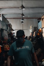 A person wearing a cap and a mask stands in a dimly lit indoor market. The market is lined with various stalls and overhead lights cast soft shadows. The person is wearing a shirt with the word 'equality' printed on it.