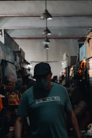 A person wearing a cap and a mask stands in a dimly lit indoor market. The market is lined with various stalls and overhead lights cast soft shadows. The person is wearing a shirt with the word 'equality' printed on it.