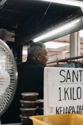 An older man is seated in a market stall, with stacks of containers filled with a brown substance, likely some type of food product. Beside him, a handwritten sign on a white board provides information about the item for sale, including 'Santan' and the price per kilo. The setting suggests a market environment with overhead lights and a visible ceiling structure.