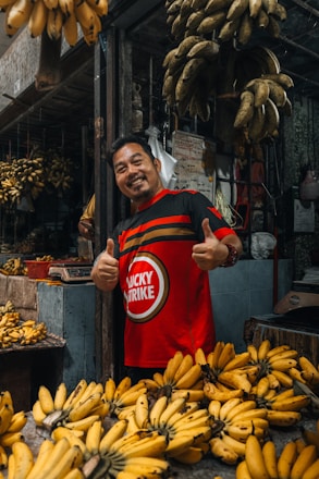 A man is smiling and giving two thumbs up while standing in a market stall filled with bunches of bananas. He is wearing a red shirt with a circular logo and the words 'Lucky Strike'. The background displays a variety of bananas, some hanging and some placed on a table, all contributing to the bustling and earthy ambiance of the market.