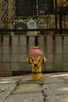 A fire hydrant painted to resemble a cartoon character with a red hat and yellow body stands on a paved sidewalk. Behind it is a stone wall and an air conditioning unit with a decorative metal fence.