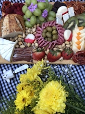 A charcuterie board featuring an assortment of foods including green grapes topped with purple flowers, sliced kiwi, various cheeses, salami, strawberries, raspberries, pistachios, bread, and olives. The spread is set on a checkered surface and accompanied by a bouquet of bright yellow flowers.