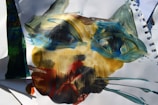 A painting featuring abstract brush strokes forming a face-like shape, with prominent use of blues, greens, yellows, and reds on white paper. The texture is rough and expressive, with overlapping colors and dynamic lines creating a sense of movement.
