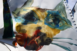 A painting featuring abstract brush strokes forming a face-like shape, with prominent use of blues, greens, yellows, and reds on white paper. The texture is rough and expressive, with overlapping colors and dynamic lines creating a sense of movement.