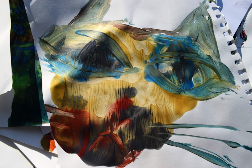 A painting featuring abstract brush strokes forming a face-like shape, with prominent use of blues, greens, yellows, and reds on white paper. The texture is rough and expressive, with overlapping colors and dynamic lines creating a sense of movement.
