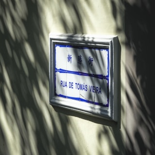 A street sign mounted on a light-colored wall with sunlight casting shadow patterns. The sign includes blue and white tiles with both Chinese characters and Portuguese text 'Rua de Tomás Vieira'.