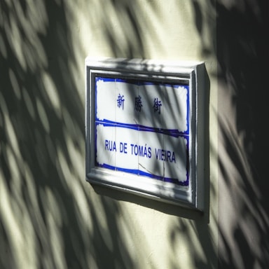 A street sign mounted on a light-colored wall with sunlight casting shadow patterns. The sign includes blue and white tiles with both Chinese characters and Portuguese text 'Rua de Tomás Vieira'.