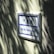 A street sign mounted on a light-colored wall with sunlight casting shadow patterns. The sign includes blue and white tiles with both Chinese characters and Portuguese text 'Rua de Tomás Vieira'.