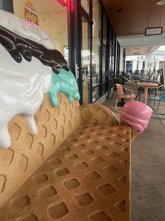 A bench designed to resemble an ice cream cone with scoops of ice cream and chocolate drizzles on top is situated outside a shop. The scene includes tables and chairs lined along a walkway, with a store entrance visible in the background, featuring signs for various establishments.
