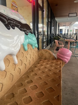A bench designed to resemble an ice cream cone with scoops of ice cream and chocolate drizzles on top is situated outside a shop. The scene includes tables and chairs lined along a walkway, with a store entrance visible in the background, featuring signs for various establishments.