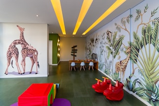 A children's playroom with vibrant, colorful wall murals featuring giraffes and jungle foliage. The room is decorated with playful elements including red toy animals and a multicolored padded cube. A table with small chairs is at the end of the room, and the ceiling has yellow recessed lights.