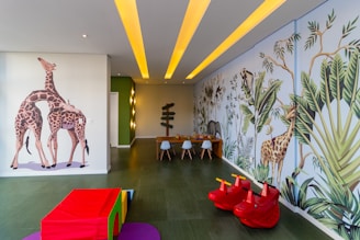 A children's playroom with vibrant, colorful wall murals featuring giraffes and jungle foliage. The room is decorated with playful elements including red toy animals and a multicolored padded cube. A table with small chairs is at the end of the room, and the ceiling has yellow recessed lights.