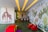A children's playroom with vibrant, colorful wall murals featuring giraffes and jungle foliage. The room is decorated with playful elements including red toy animals and a multicolored padded cube. A table with small chairs is at the end of the room, and the ceiling has yellow recessed lights.