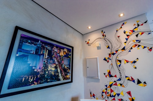 A framed picture of a vibrant urban cityscape is mounted on a white wall, showcasing a bustling city at night with brightly lit buildings and streets. To the right, the wall features an abstract mural incorporating swirling black lines and geometric shapes in bold colors such as red, blue, yellow, and black, forming a tree-like design.