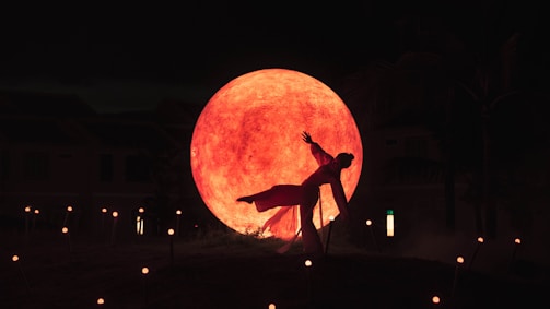 A dancer poses dramatically in front of a large, illuminated orange-red sphere, which resembles the moon. The scene is set in a dimly lit outdoor environment with scattered small lights on the ground, adding to the mystical atmosphere.