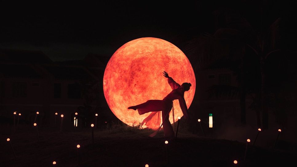 A dancer poses dramatically in front of a large, illuminated orange-red sphere, which resembles the moon. The scene is set in a dimly lit outdoor environment with scattered small lights on the ground, adding to the mystical atmosphere.