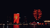 Nighttime cityscape featuring a building illuminated with a large Vietnamese flag. Numerous red lanterns, arranged in the shape of a heart, float in the sky. A bridge with red lighting spans across a body of water reflecting the lights.