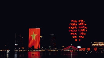 Nighttime cityscape featuring a building illuminated with a large Vietnamese flag. Numerous red lanterns, arranged in the shape of a heart, float in the sky. A bridge with red lighting spans across a body of water reflecting the lights.
