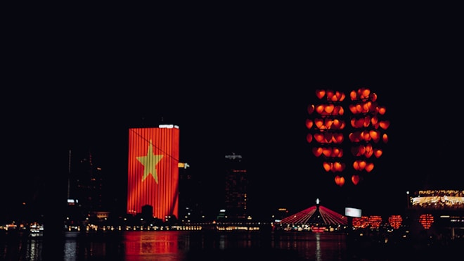 Nighttime cityscape featuring a building illuminated with a large Vietnamese flag. Numerous red lanterns, arranged in the shape of a heart, float in the sky. A bridge with red lighting spans across a body of water reflecting the lights.